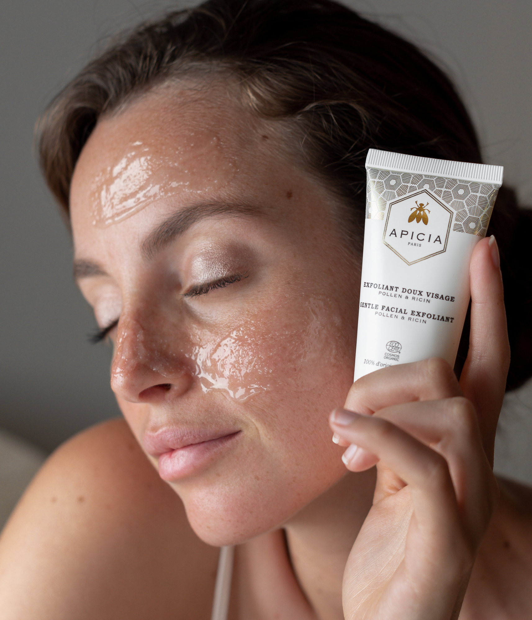 Fille tenant un tube d'exfoliant blanc et doré près de son visage, recouvert de l'exfoliant transparent et brillant.