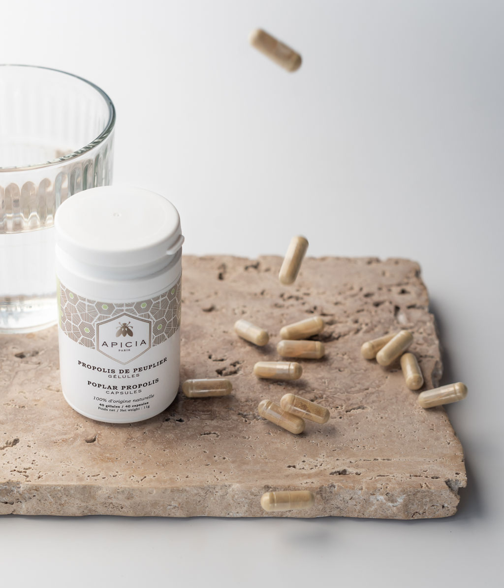 Flacon blanc de propolis à côté d'un verre d'eau et de gélules beige qui tombent.
