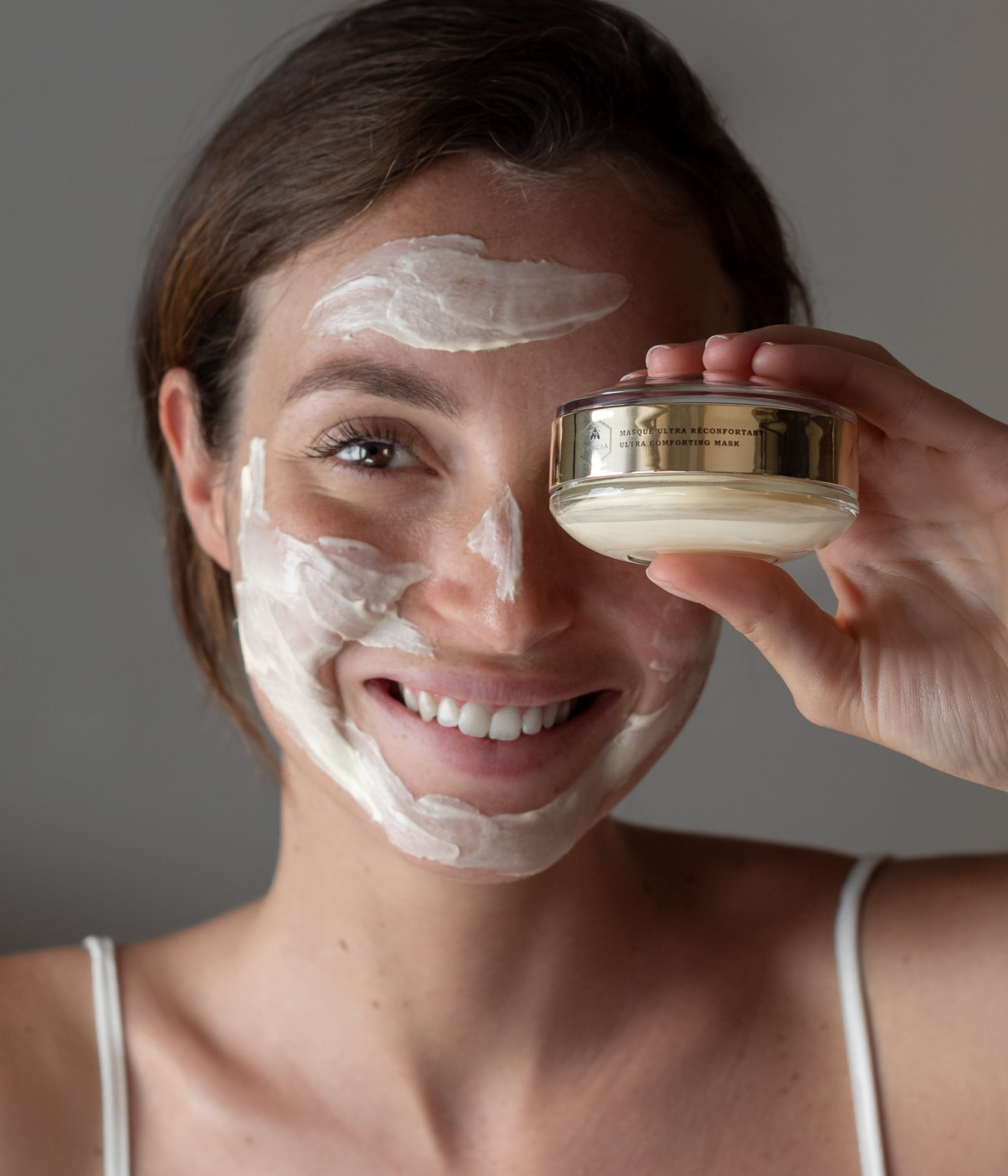 Femme tenant un flacon oval doré en verre devant son œil gauche, avec un masque blanc posé sur son visage.