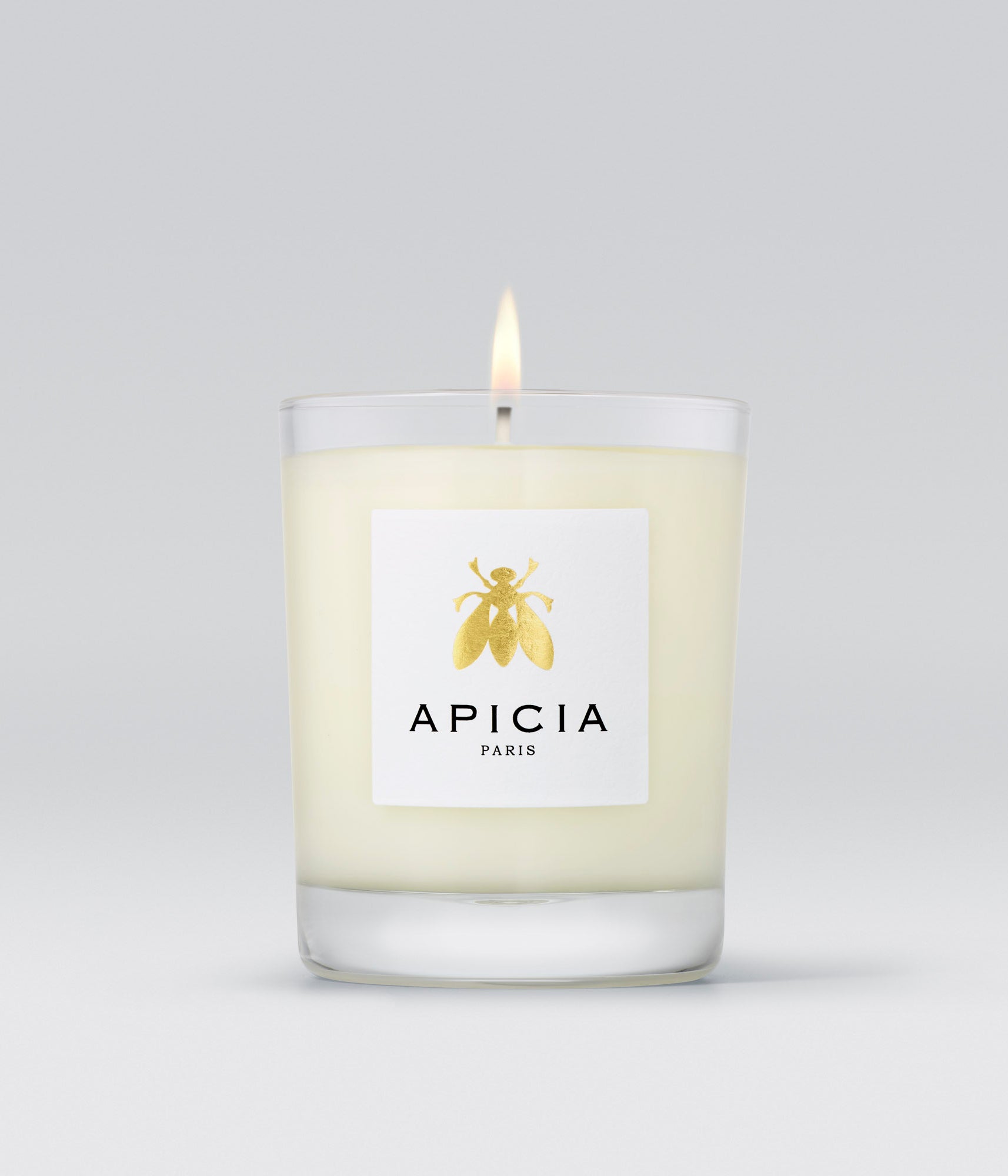 Packshot d'une bougie blanche dans un verre transparent, avec une étiquette blanche comportant une abeille dorée.