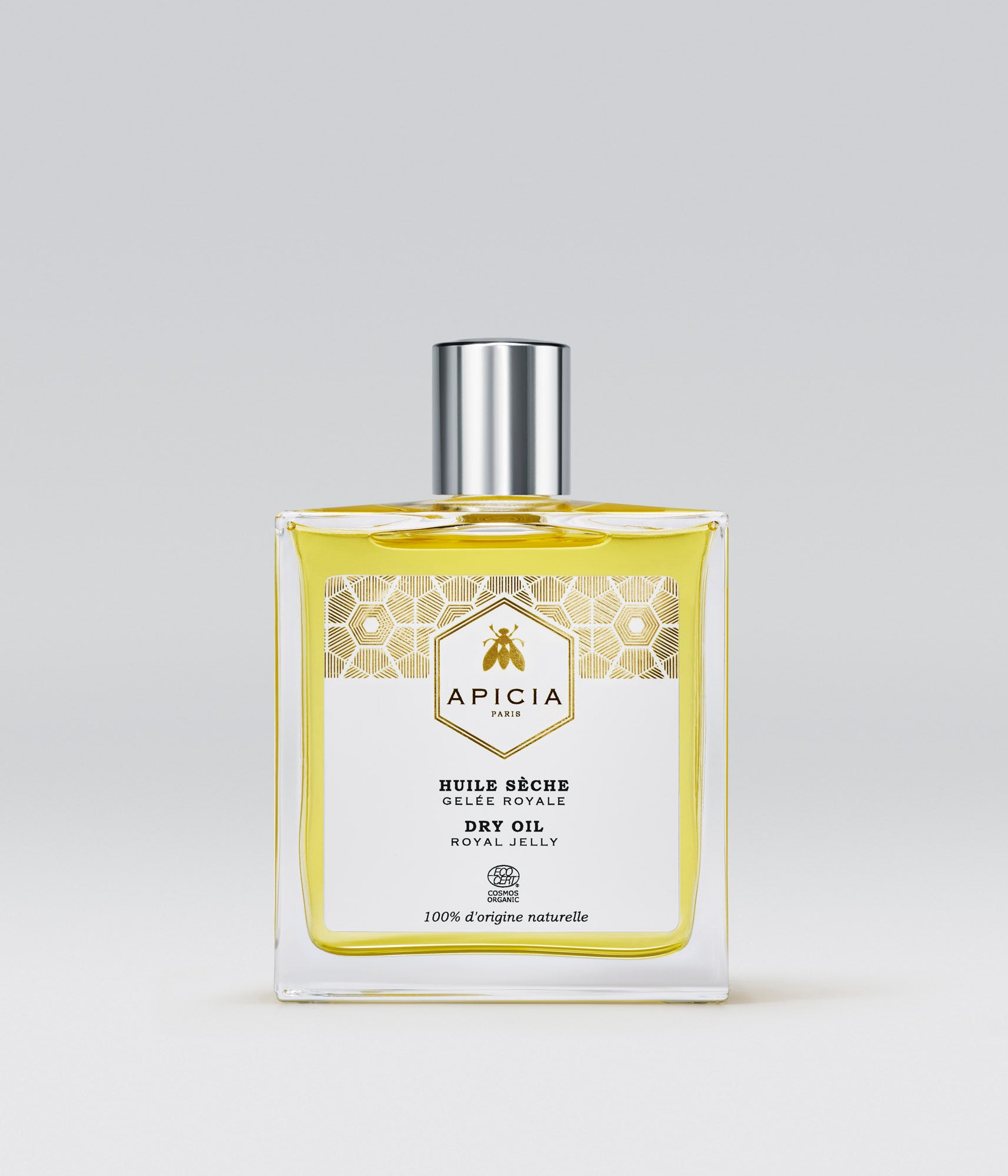 Packshot d'un flacon en verre transparent rectangulaire contenant une huile jaune, sur fond gris.