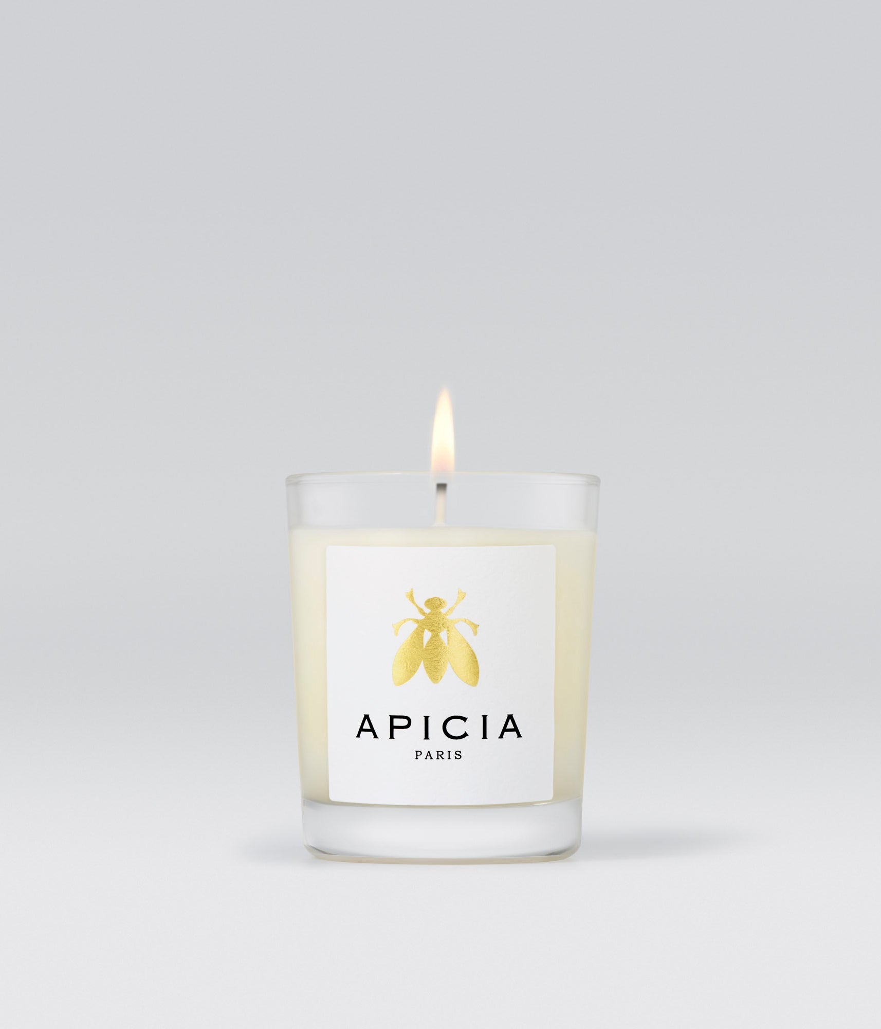 Packshot d’une petite bougie blanche dans un verre transparent et une étiquette blanche avec une abeille dorée.