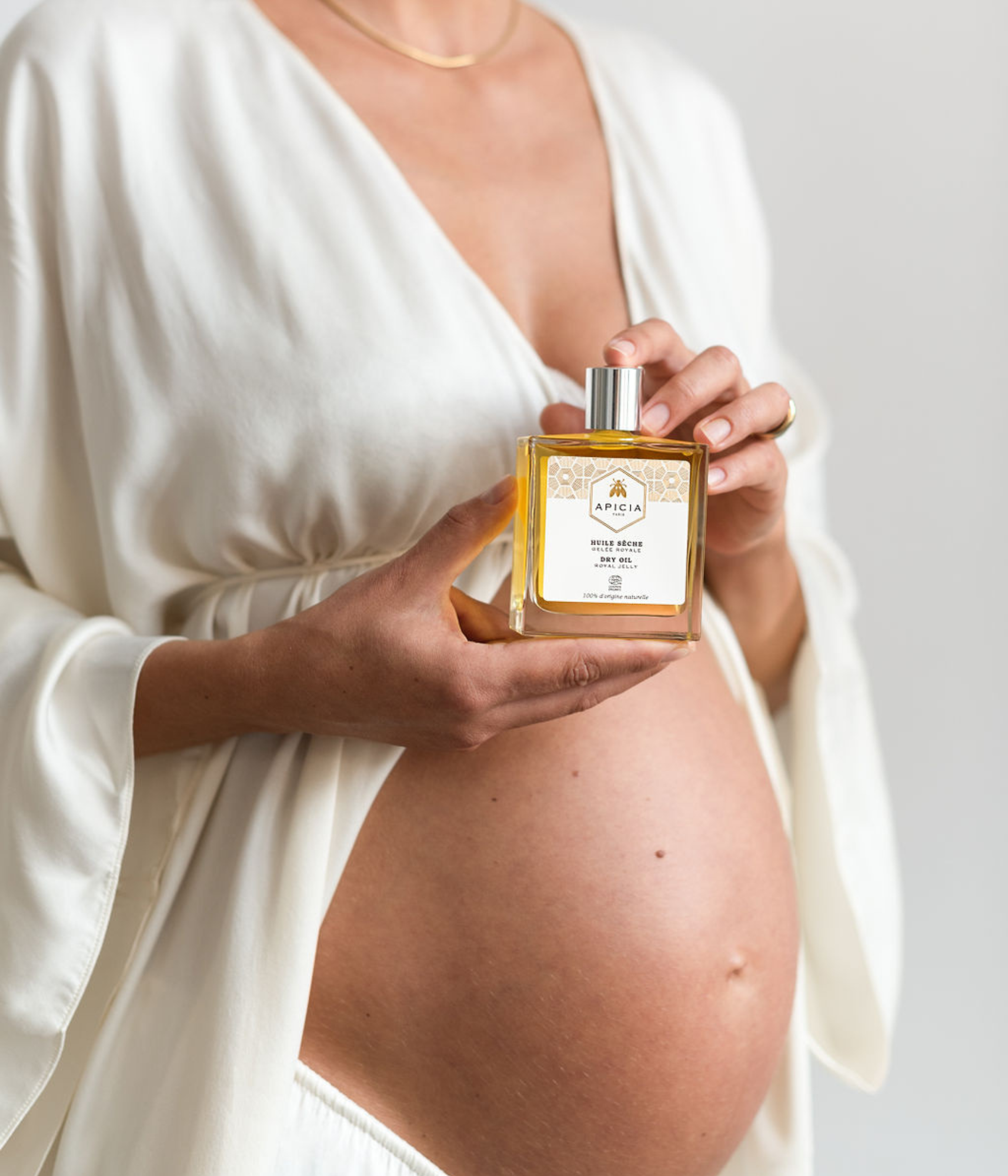 Femme enceinte tenant un flacon rectangulaire en verre transparent contenant de l’huile dans ses deux mains qui reposent près de son ventre.