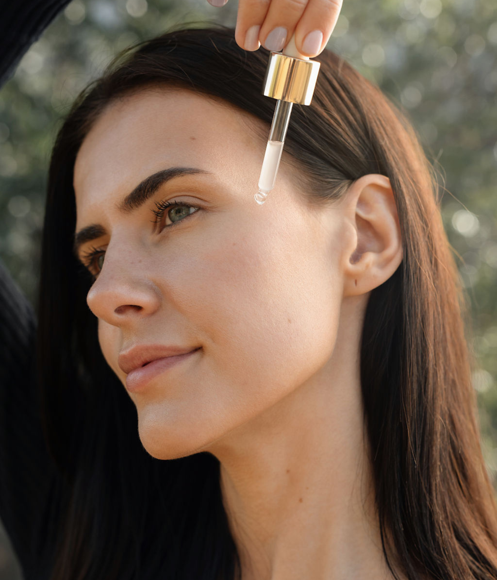 Femme aux cheveux bruns de profil, tenant une pipette avec un liquide transparent sur le côté de son visage.